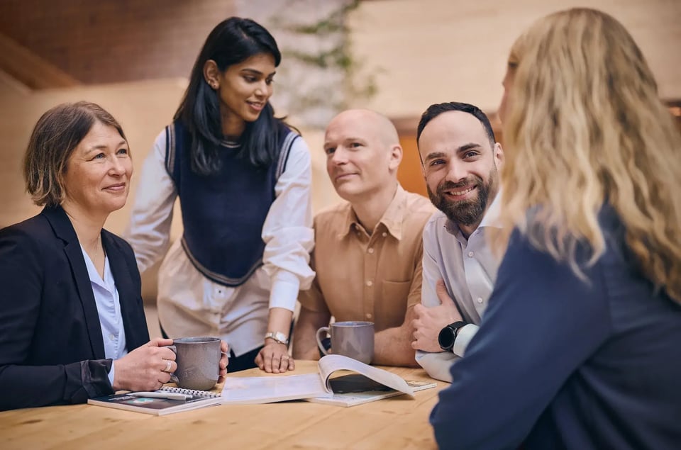 Pharmetheus employees Malin G, Mounika S, Jakob R, and Rami A gathered around a wooden table for a collaborative team meeting in a modern office space.